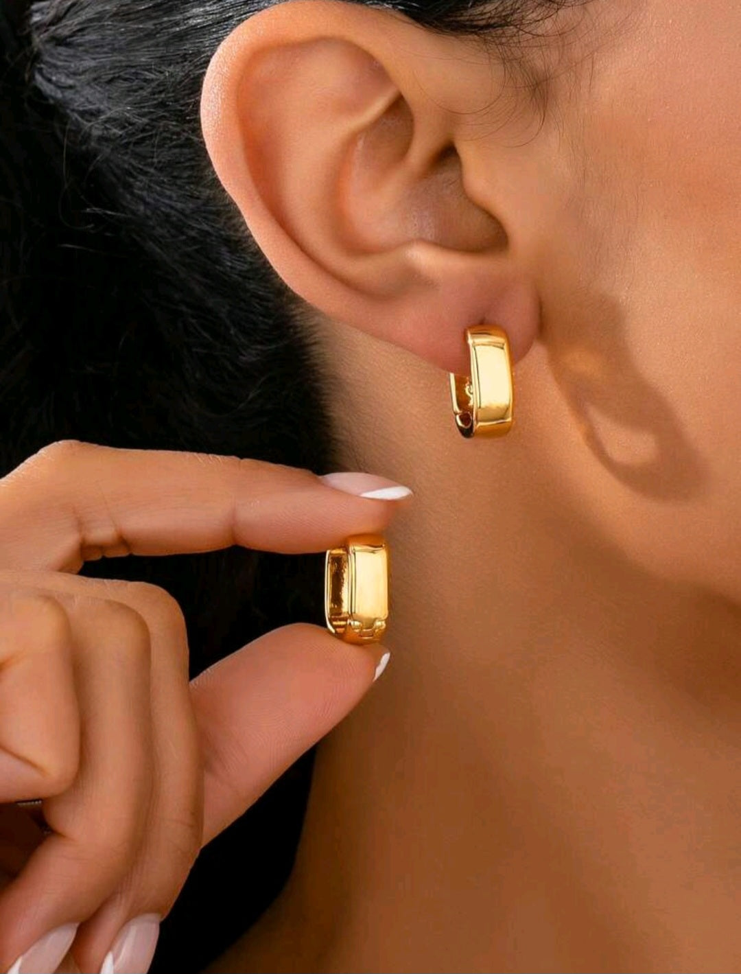 Gold hoop earrings being worn and held by a hand against a neutral background